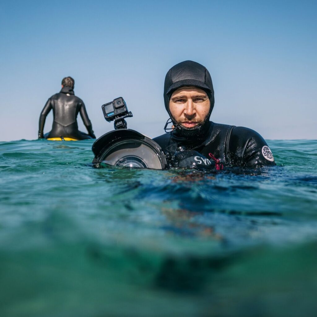 Photographer in the ocean with man sitting on surfboard behind